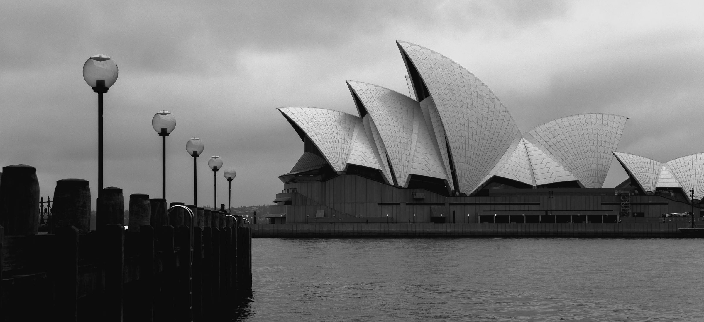 The Sydney Opera House under heavy cloud, seen across the harbour past timber wharf piles